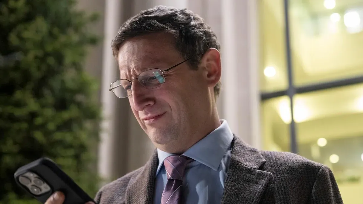 Tim Robinson in episode 3 of The Chair Company. He is wearing glasses and a gray suit, looking at his phone.